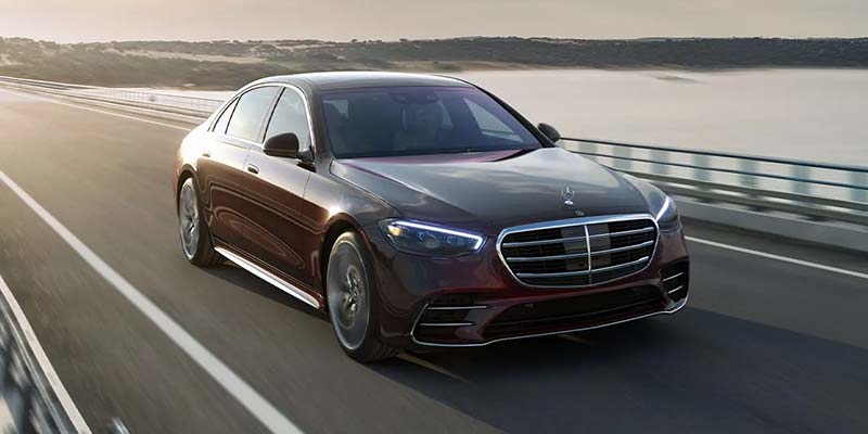 A dark Mercedes-Benz sedan drives along a coastal road beside the water.